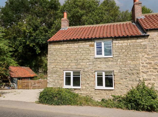A stone cottage with red tile roof and white framed windows next to a small gravel area with a wooden fence and surrounded by greenery at Yon Cottage in Stonegrave near Hovingham