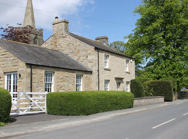 Stone house with white framed windows and doors surrounded by green hedges next to a road and a church tower in the background at Vine Cottage in Pilling
