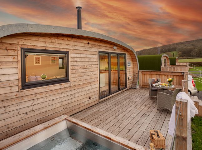 A wooden cabin with a deck patio area featuring a hot tub a round table with chairs and a scenic view at Middle Longlands in Cartmel