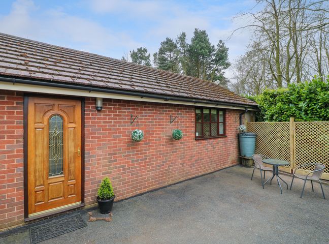 A brick house exterior with a wooden door a window hanging plants a rain barrel and a small table with two chairs at Woodstock Lodge in Hednesford