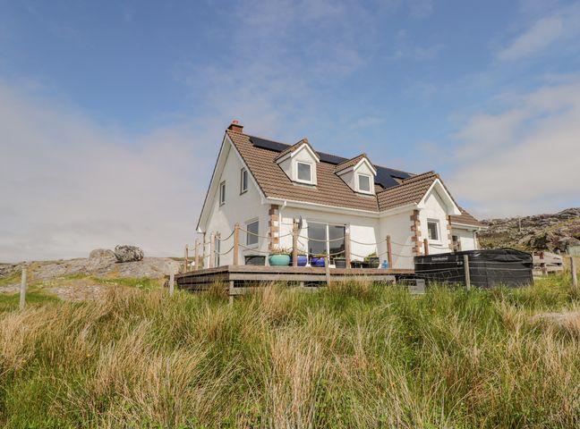A white house with a deck and solar panels on the roof surrounded by grass and rocks at Beachview in Oldshore Beg near Kinlochbervie