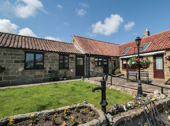 A courtyard with grass and flower beds surrounded by stone buildings with tiled roofs at Beaconsfield Cottage Burniston near Scalby