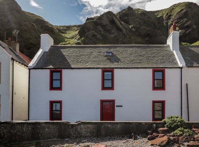 A white two-story house with red door and window frames against a rocky hill at The Laurels in Pennan near Gardenstown
