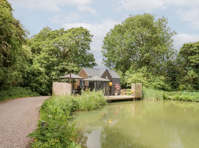 An outdoor area with a house by a pond at Maple Lodge at Bridge Lake Farm & Fishery Chacombe
