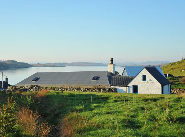 A white house with a gray roof near a body of water with hills in the background at Tigh Bhisa Blackhouse in Tolsta Chaolais