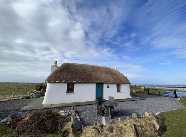 A white thatched cottage with a blue door and two windows surrounded by gravel and grass at Cuir na Bhoir in Creagorry