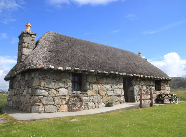 A stone cottage with thatched roof and wooden wheels outside on grass at Smiddy Cottage in Smercleit near Daliburgh