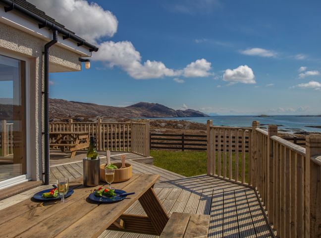 A wooden deck with a picnic table set with plates and glasses overlooking a rocky shoreline and sea at Rockpool in Glendale Isle of South Uist