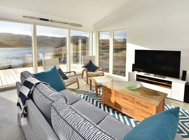 A living room with a striped sofa blue cushions wooden coffee table armchairs and a large TV with glass doors overlooking a lake at Beach Cottage in Dalbeg