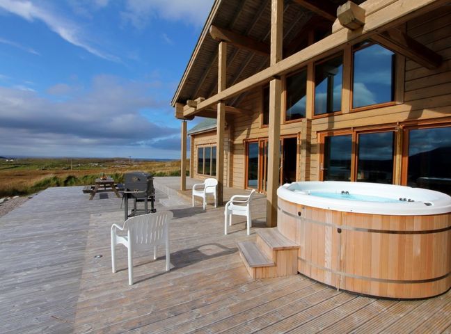 An outdoor wooden deck with a hot tub white plastic chairs a BBQ grill and a picnic table at Eagle Lodge in Uig near Stornoway
