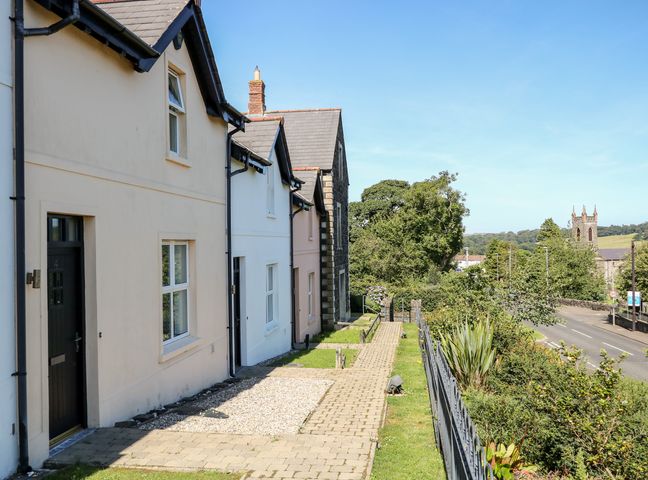 A row of houses with a paved walkway and a garden area next to a road with trees and a stone tower in the distance at 4 The Cloisters in Bushmills County Antrim