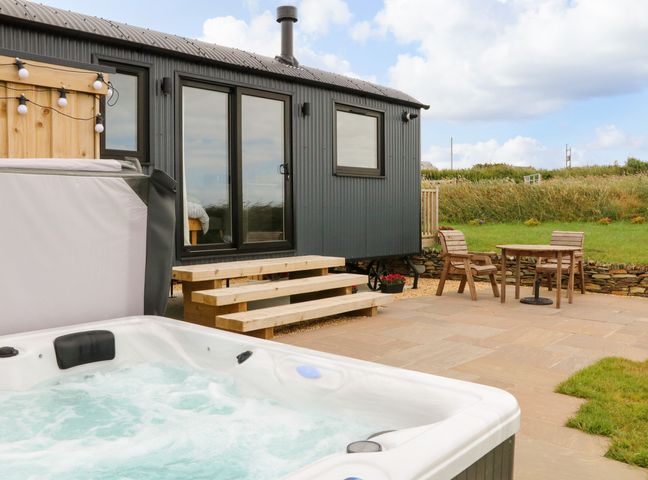 An outdoor patio area with a hot tub wooden steps leading to a grey building and a wooden table with two chairs on a stone patio at Northcott Hut in Stibb near Bude