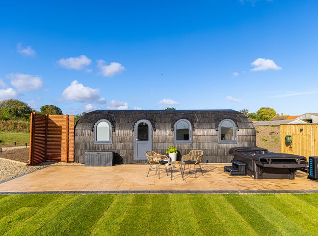 A small curved wooden building with three windows and a door with outdoor chairs a table and a hot tub on a patio at Veiko - Mayflower Meadow in Burshill near Brandesburton