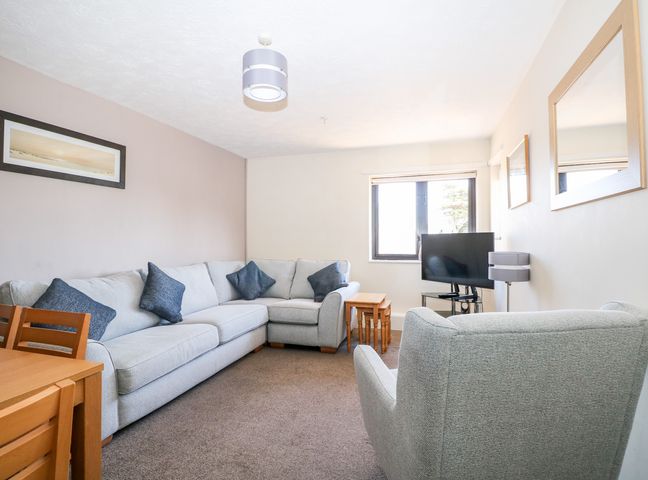 A living room with a grey sectional sofa blue cushions a wooden dining table a TV on a stand next to a window and wall mirrors at Flat 34 - Marine Court in Littlehampton