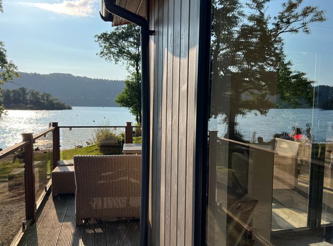 A lakeside deck with wicker chairs and a glass railing at Loubi's Lakeside Lodge M1 Fallbarrow Park near Bowness-On-Windermere