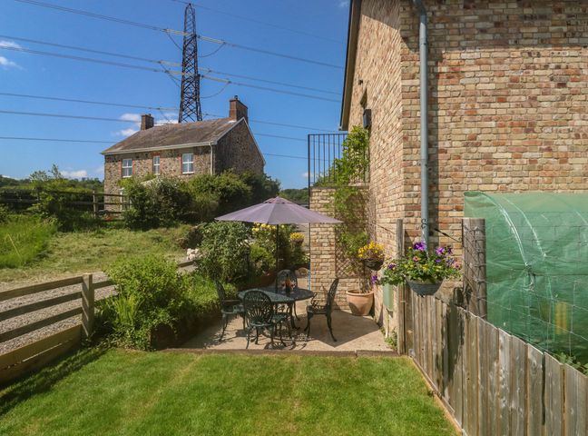 An outdoor patio with a table umbrella and chairs next to a brick building and garden at The Mews in Bovey Tracey near Chudleigh Knighton