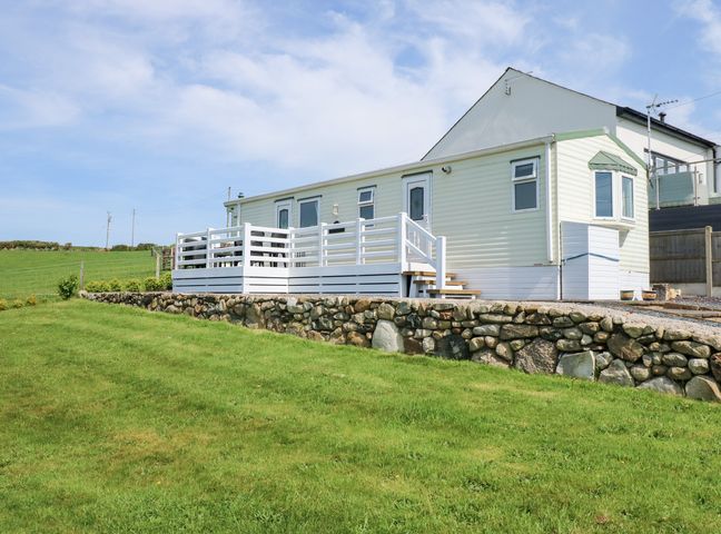 An exterior view of a single-story caravan with a white deck and stone retaining wall on a grassy landscape at Ty Newydd Caravan in Llannor near Pwllheli