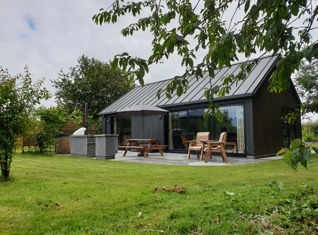 A garden with a small modern building with a metal roof wooden chairs a picnic table with umbrella and an outdoor pizza oven at Blackcap in Huggate near Bishop Wilton