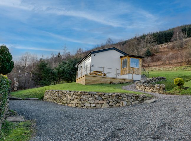 A small house with a wooden porch and stone retaining walls on a grassy hill at Ciaran Cottage in Fearnan near Kenmore