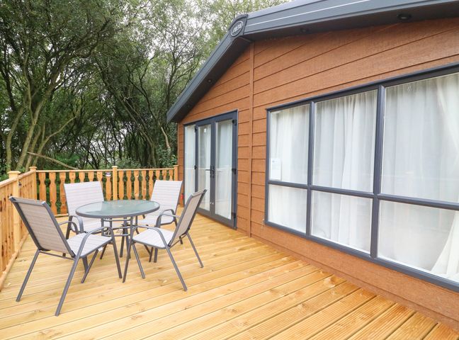 A wooden deck with a round glass table and four chairs outside a building with large windows at Barneys Retreat in Carnforth