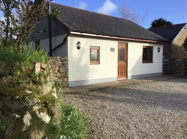 A small house with two windows and a door in a gravel yard with a stone wall and plants at Tregonning Lodge in Ashton near Rosudgeon