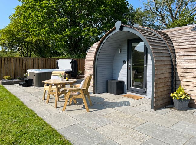 A wooden pod with a glass door on a stone patio with a hot tub wooden table and chairs and a flower pot at Y Dderwen in Bontuchel near Ruthin