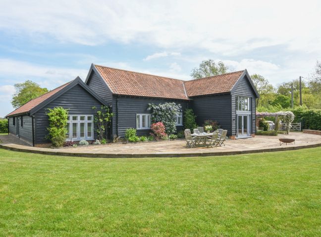 A house with dark wooden exterior and tiled roof surrounded by a lawn and outdoor patio with table and chairs at Looms Barn Hasketon Woodbridge