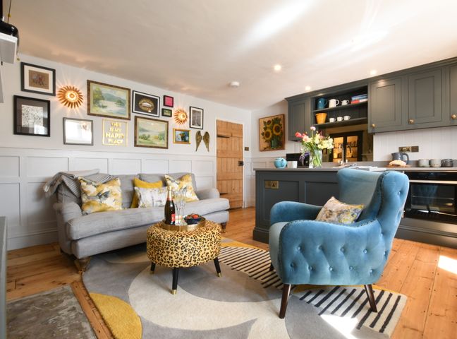 A living room with a blue armchair a leopard print ottoman a gray sofa and a kitchen area at Cragside Aldeburgh in Aldeburgh
