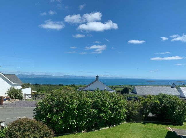 A view of houses with green bushes and a lawn overlooking the sea under a blue sky at Watlands in Mynytho near Abersoch