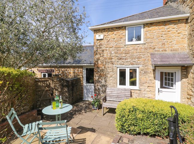 A small stone patio with a round table and chairs next to a wooden bench outside a stone house at Tiddlers Cottage in Uploders near Bridport