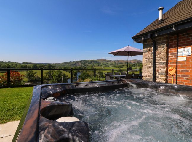 An outdoor hot tub next to a stone and brick building with a table, chairs, and umbrella overlooking green fields and hills at Pen Y Banc in Llanfaredd near Builth Wells