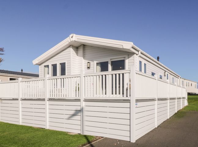 A white mobile home with a white fenced deck on a grassy area at Dershel Lodge in Southerness