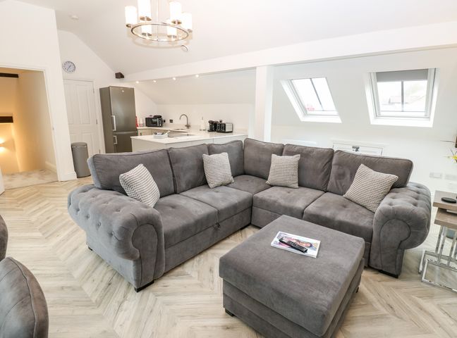 A living room with a grey sectional sofa and ottoman next to a kitchen with a refrigerator and sink at The Penthouse in Boroughbridge