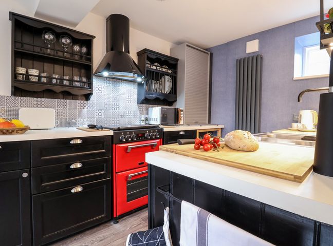 A kitchen with black cabinets a red oven a wooden cutting board with bread and tomatoes and a tiled backsplash at Tower End Cottage in Melton