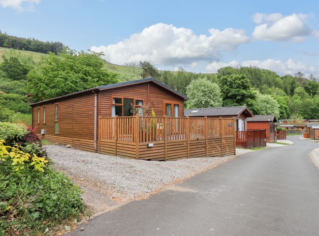A wooden lodge with a fenced deck next to a paved road with other wooden buildings and trees in the background at Hollytree Lodge Beckside 1 Limefitt Park near Windermere