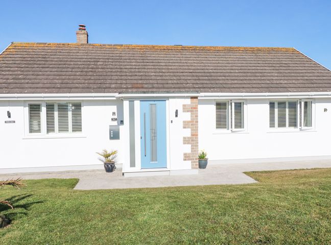 A white bungalow with a blue front door and three windows on a sunny day at Palm View in Holywell Bay