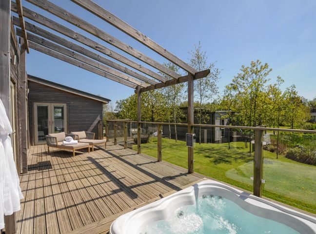 A wooden deck with a hot tub a lounge chair and a wooden pergola overlooking a garden at 11 Meadow Retreat in Dobwalls