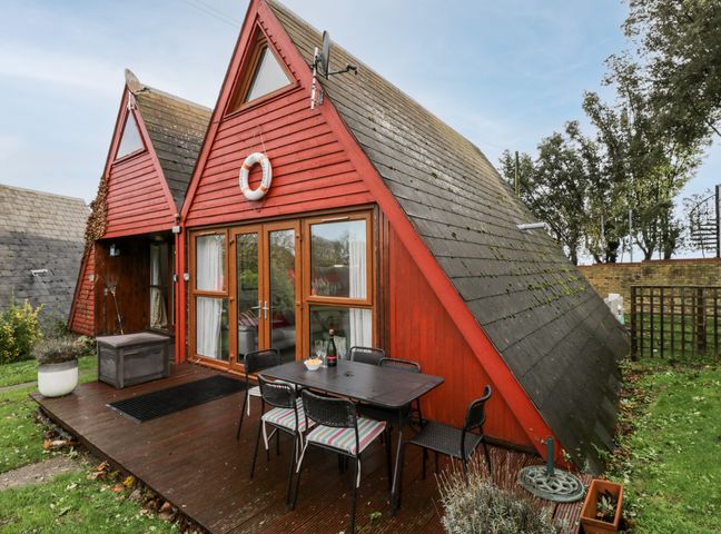 A red wooden cabin with triangular roof and outdoor dining table on wooden deck at Beachmaster in Kingsdown