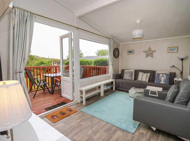 A living room with a gray sofa and armchair a blue rug and a door opening onto a wooden deck with black chairs and a table at Chalet 64 in Tywyn