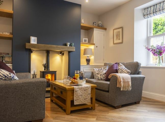 A living room with two gray sofas a wooden coffee table with breakfast items a fireplace and a window with flowers at Wisteria Cottage in Warkworth