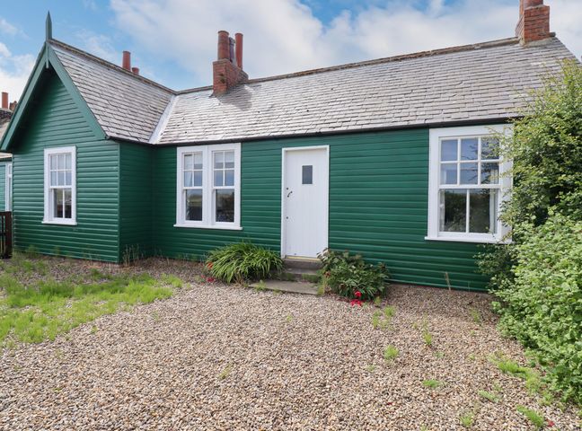 An exterior view of a green wooden house with white windows and a white door with a gravel yard at Sand Dune Cottage in Bamburgh