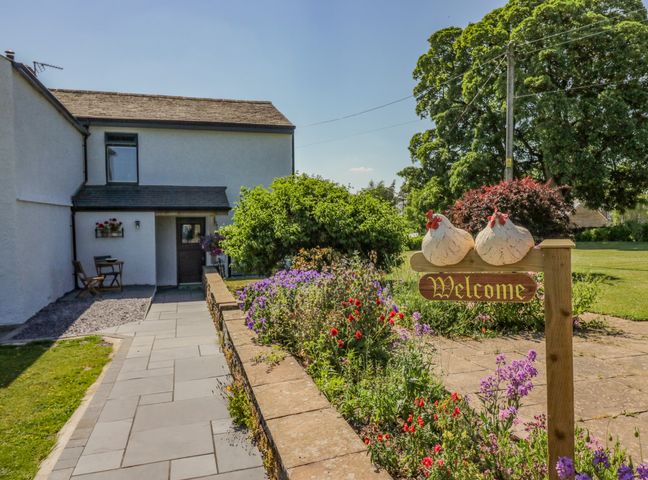 A paved walkway leading to a house with a garden bed of flowers and a welcome sign with two decorative chickens at How Hill Farm Cottage in Greystoke