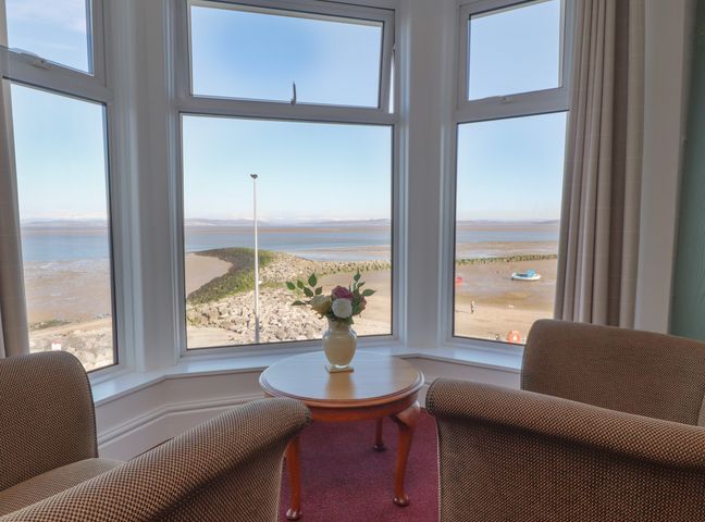 A sitting area with two armchairs and a round wooden table with a vase of flowers by bay windows overlooking a beach and water at Eden Bay Retreat in Morecambe