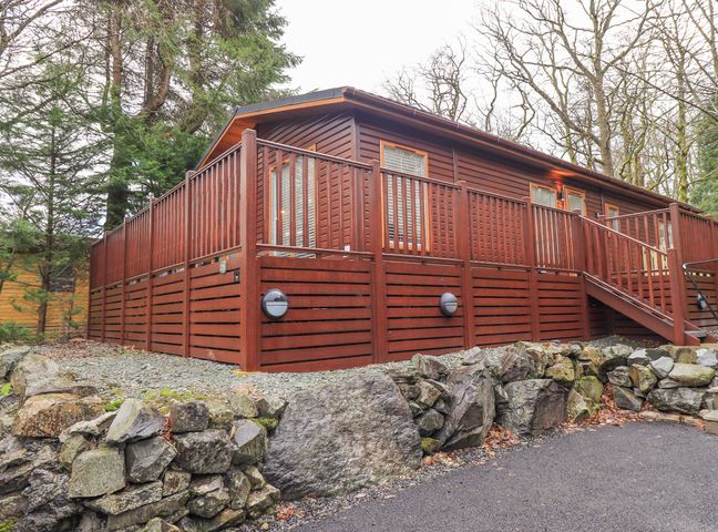 A wooden cabin with a raised deck and stairs surrounded by trees and a stone wall at 25 Thirlmere in Troutbeck Bridge