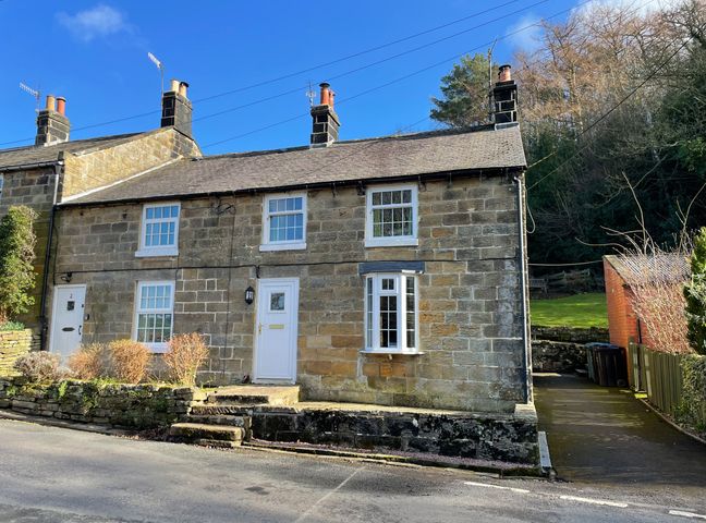 A stone cottage with white framed windows and doors next to a driveway and greenery at The Old Sweet Shop in Grosmont