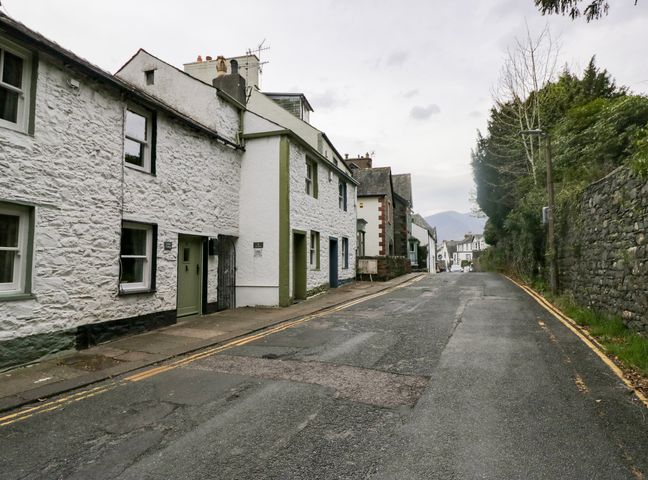A narrow street with stone houses on the left and a stone wall with trees on the right at Latch Cottage in Keswick