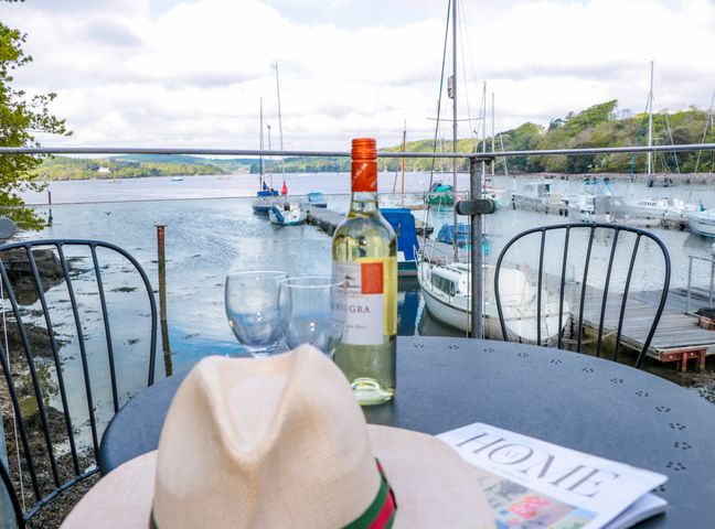 A balcony with a table holding a bottle of white wine two glasses a hat and a magazine overlooking boats docked at a marina with water and trees in the background at Spanish Boathouse in Galmpton Torbay