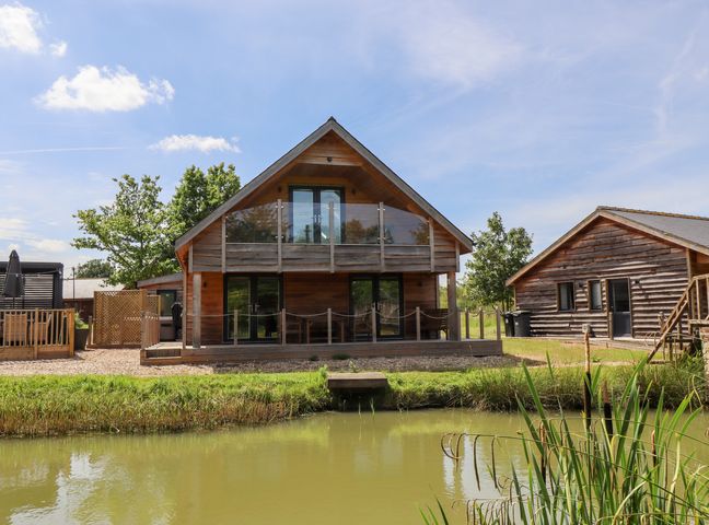 A wooden lodge with balcony and glass doors next to a pond and grassy area at Lodge 24 in South Hykeham