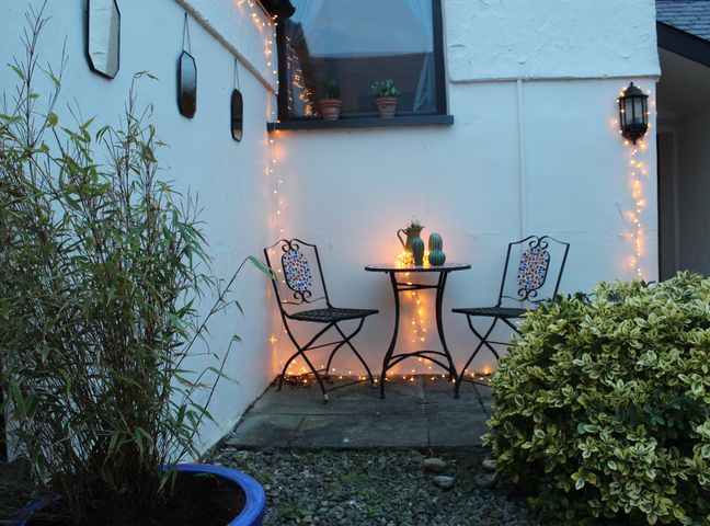 An outdoor seating area with two metal chairs and a small round table with lights stringed on the wall and table at Capel Zion Burwen near Amlwch