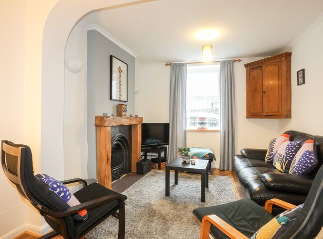 A living room with a black leather sofa and chairs a wooden fireplace a small black coffee table and a window with grey curtains at 5 Thomas Street in Llanberis